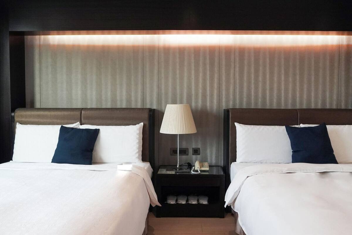 A hotel room with two beds, a nightstand with a lamp, and a wall-mounted phone.