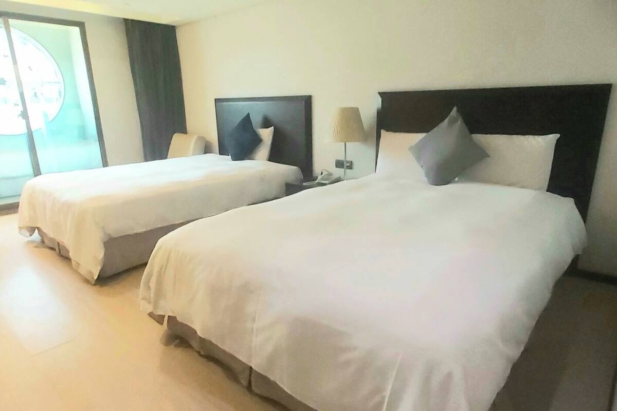 Two single beds with white linens, a dark headboard, a bedside table with a lamp, and a large window with blinds.