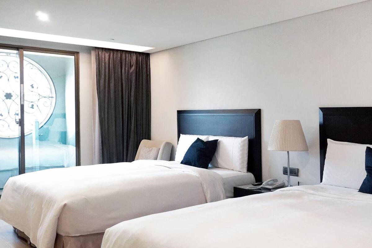 A hotel room with two beds, a large window with curtains, a bedside table with a lamp, and a wall clock.