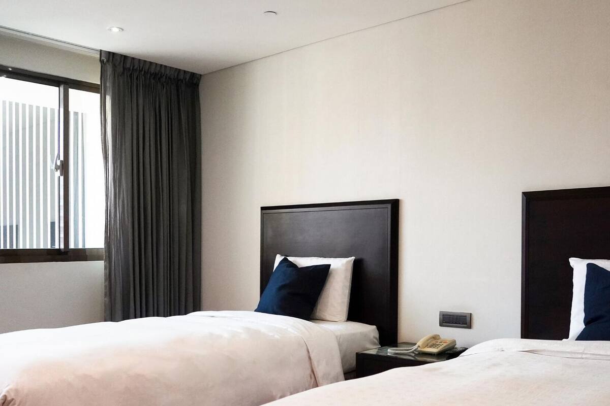 A hotel room with two beds, a window with curtains, and a bedside table with a phone.