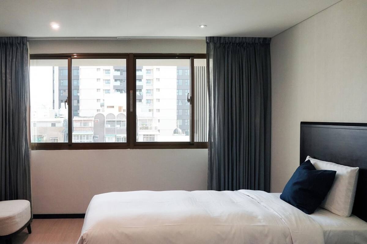 A hotel room with a large window, a bed with white bedding, a chair, and a dark headboard.