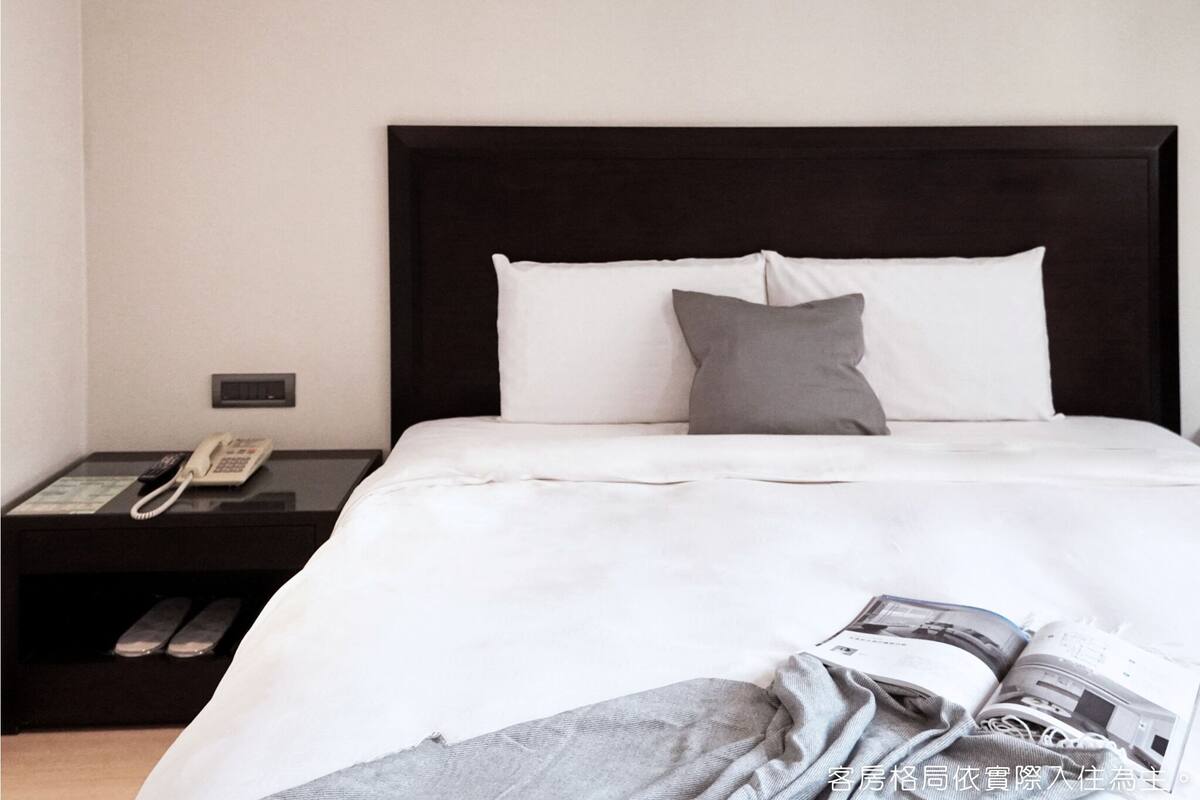 A neatly made bed with white and gray bedding, a bedside phone, and a magazine on the bed.