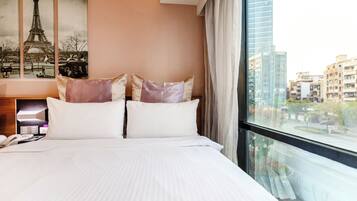 A hotel room with a large bed, a telephone, and a nightstand. There is a window with a view of a cityscape.