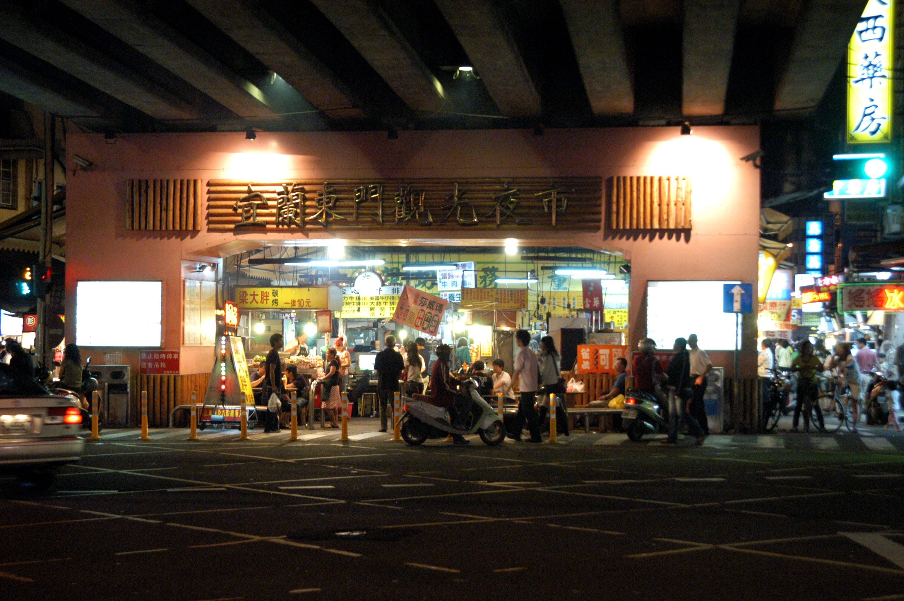 A busy street scene with a restaurant entrance, people walking, and scooters parked outside.