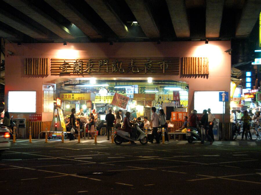 A busy street scene with a restaurant entrance, people walking, and scooters parked outside.