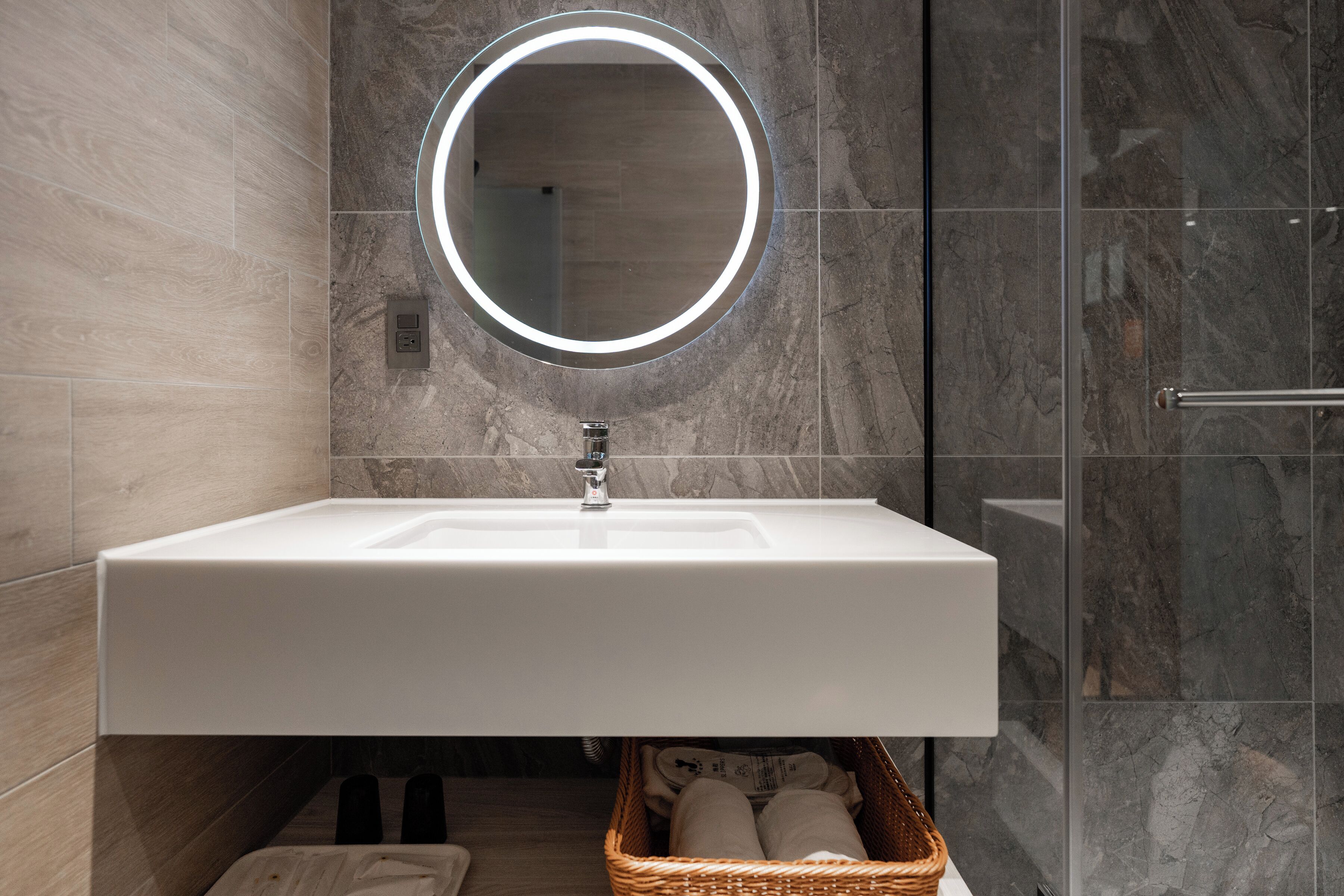 Een moderne badkamer met een witte wastafel, een ronde spiegel met verlichting, en een rieten mandje onder de wastafel.