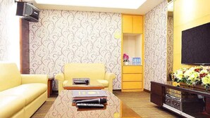 Un salon avec un canapé jaune, un fauteuil beige, une table basse avec des livres et une télécommande, un téléviseur posé sur un meuble et un papier peint à motifs.