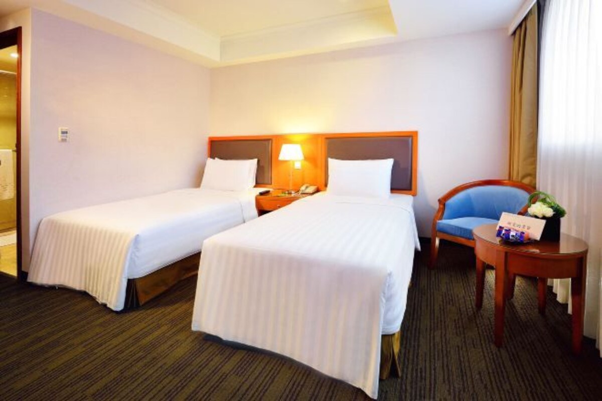 A hotel room with two beds, a nightstand with a lamp, a chair, and a small table with a vase.