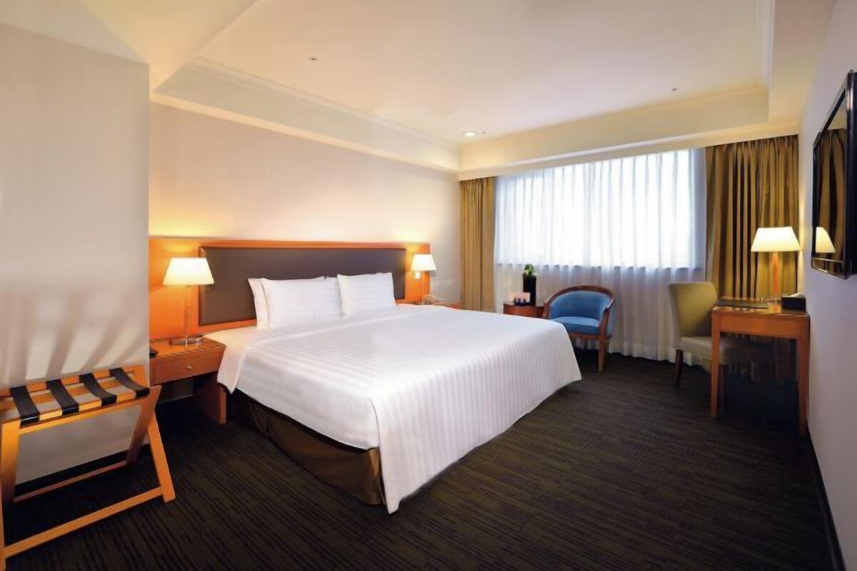 Una habitación de hotel con una cama grande, dos mesitas de noche con lámparas, una silla, un escritorio y un gran ventanal con cortinas.