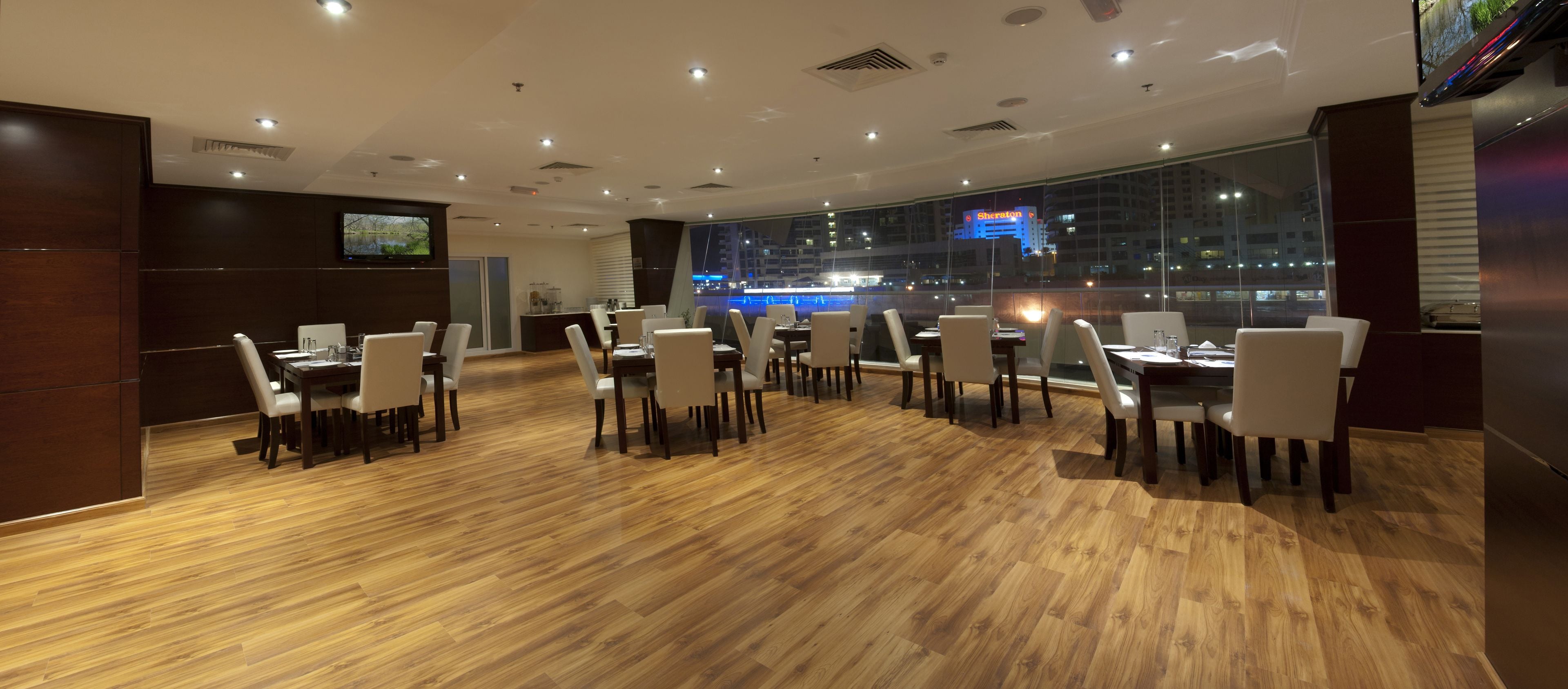 A modern dining area with wooden flooring, white chairs, and a large window offering a city view.