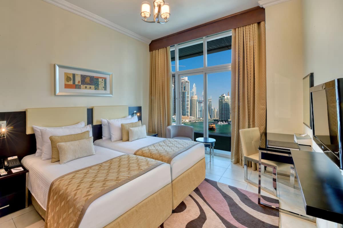 Ein Hotelzimmer mit zwei Betten, einem Schreibtisch, einem Sessel, einem Fernseher und einem großen Fenster mit Blick auf die Stadt.