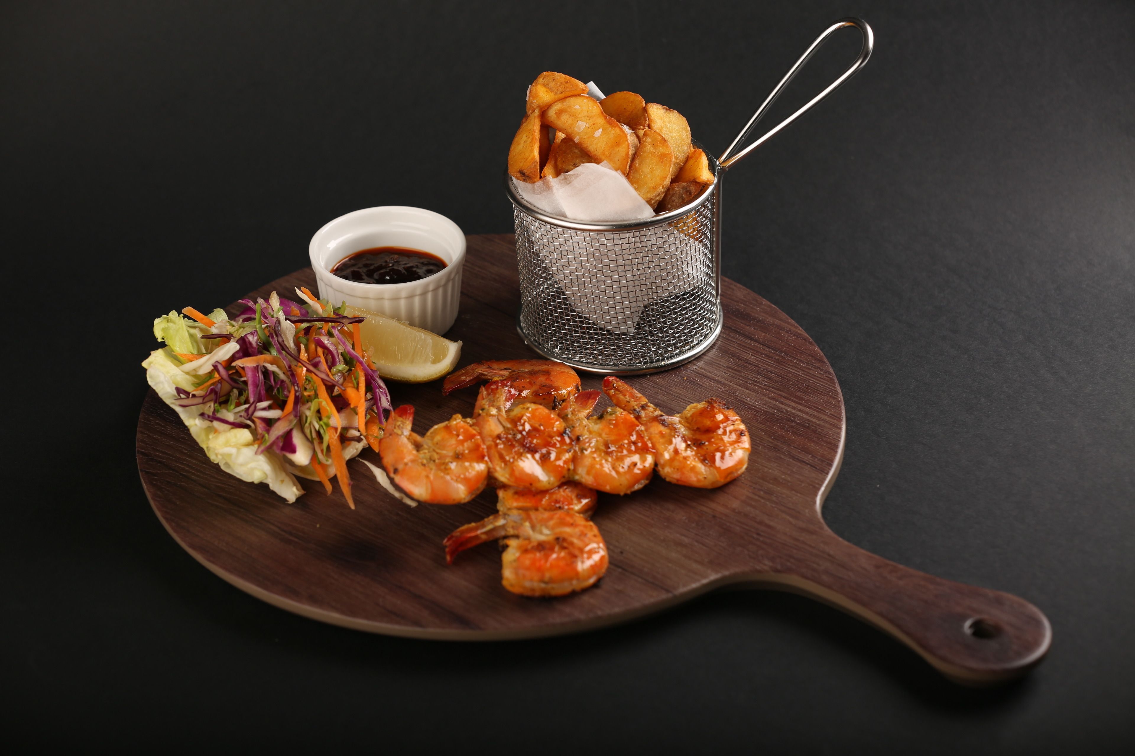 Una tabla de madera con camarones a la parrilla, una ensalada de col y un recipiente pequeño para salsa.