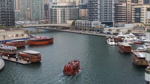 Un río con varios barcos, incluyendo un autobús turístico rojo de dos pisos y un bote marrón con cubierta de madera, rodeado de modernos edificios altos.
