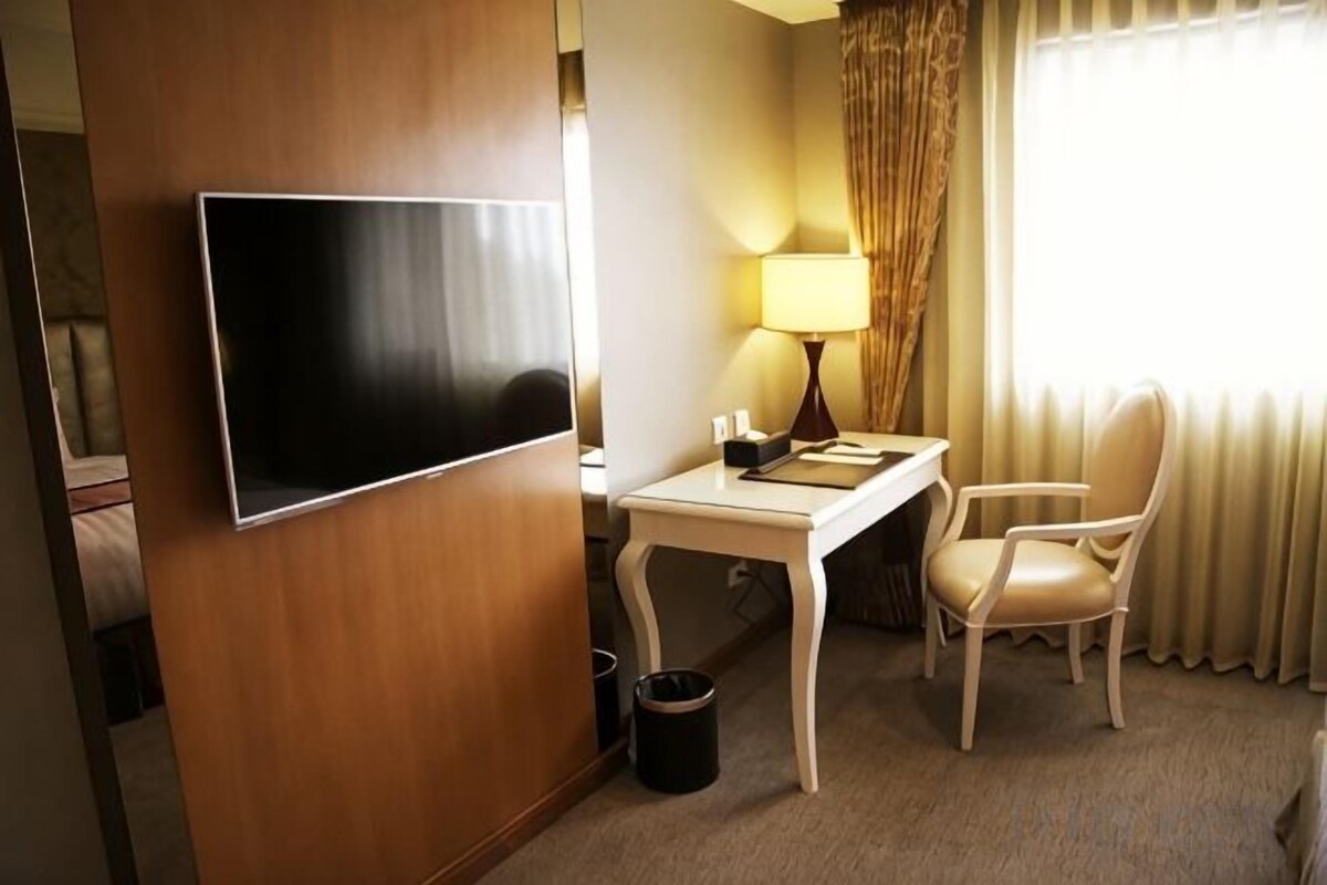 Habitación de hotel con una televisión de pantalla plana montada en la pared, un escritorio blanco pequeño con una lámpara, una silla y una ventana con cortinas.