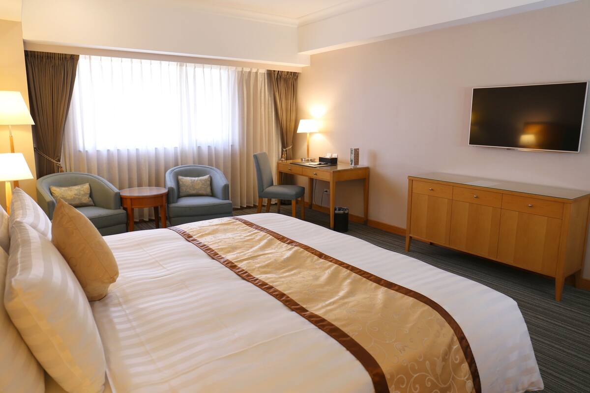 A hotel room with a large bed, two armchairs, a small table, a desk, a television, and a wardrobe.