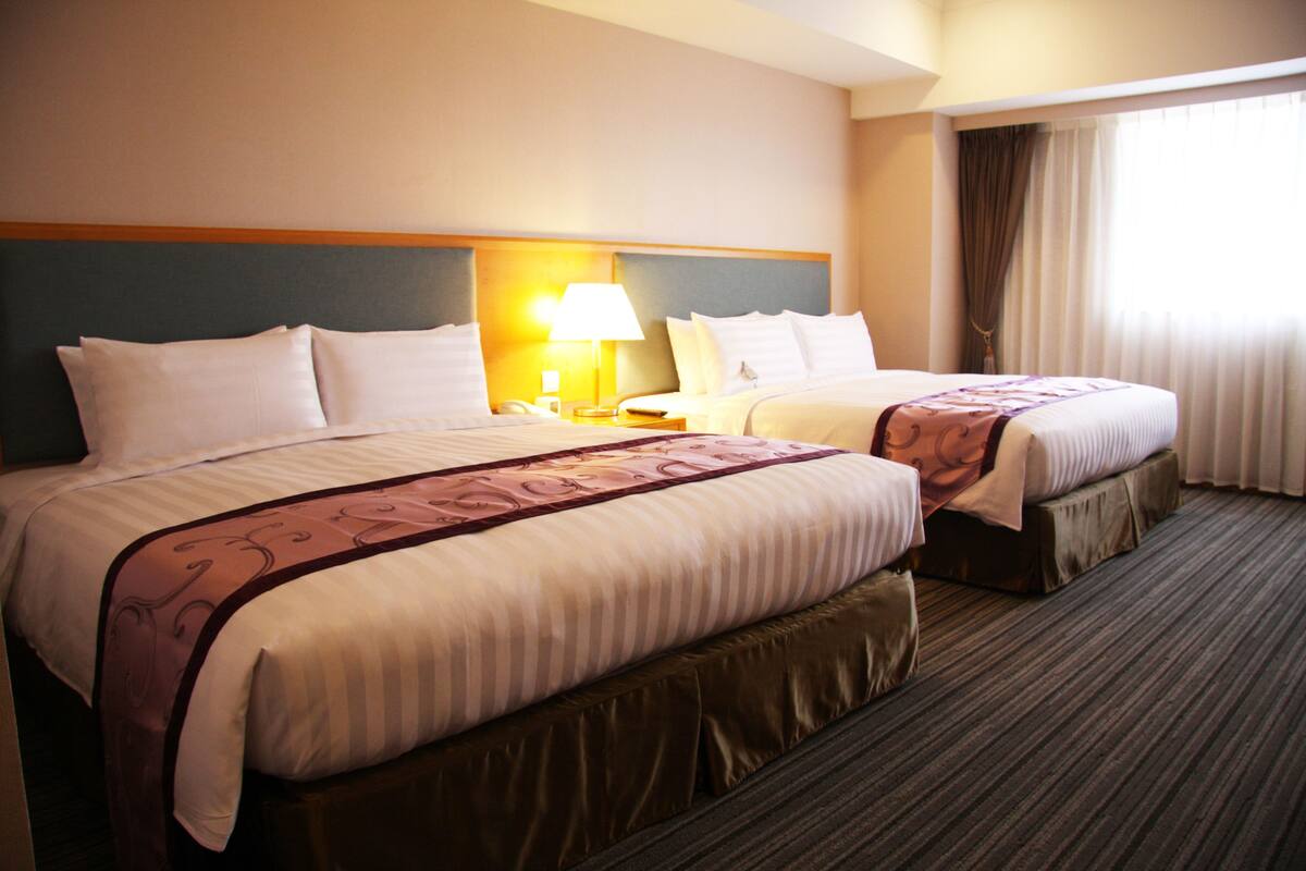 A hotel room with two beds, a nightstand, a lamp, and a window with curtains.