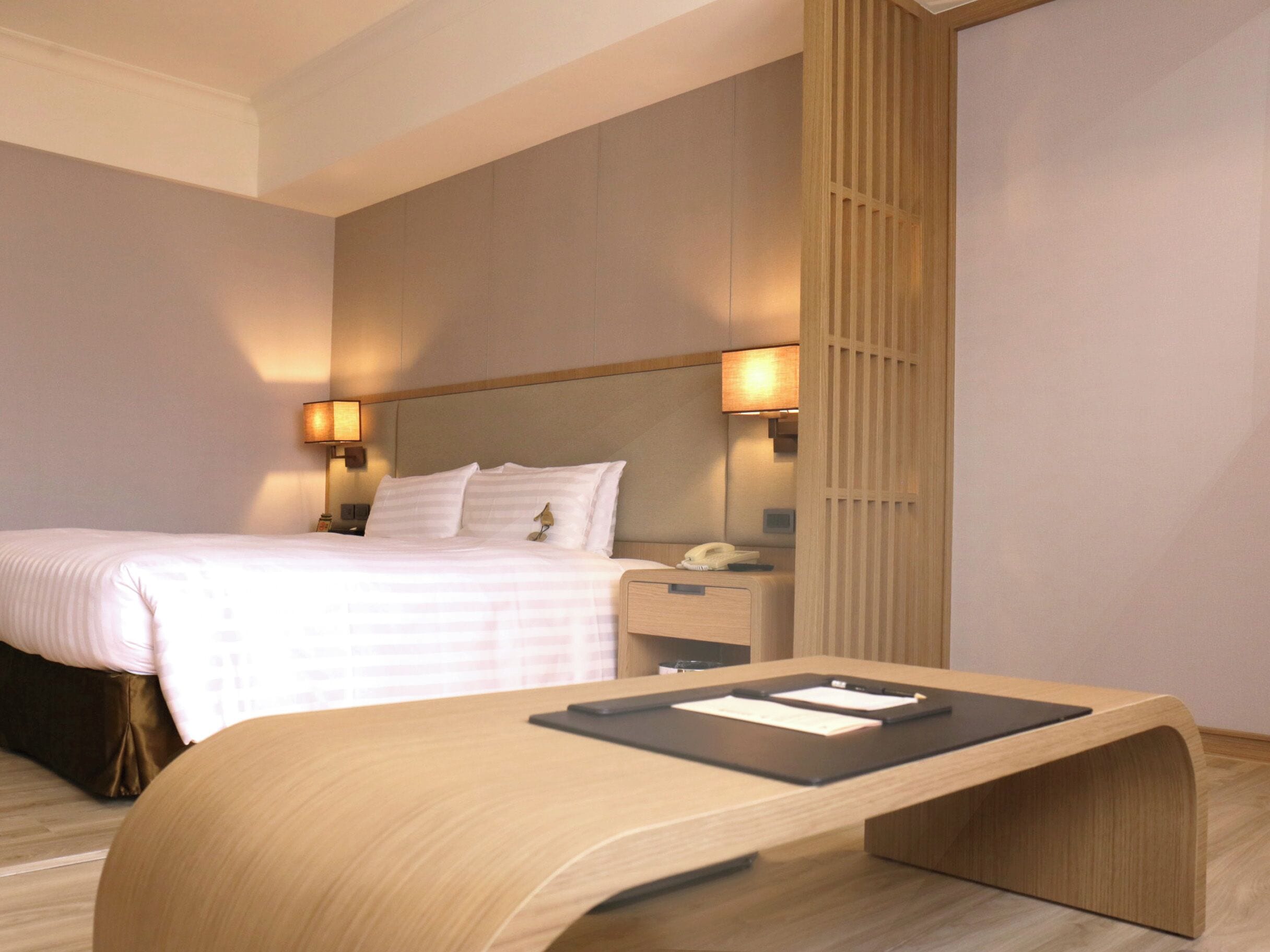 A modern hotel room with a neatly made bed, bedside lamps, a wooden desk with a laptop, and a nightstand with a phone.