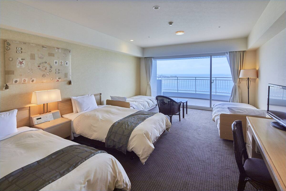 A hotel room with two beds, a desk, a chair, a lamp, and a large window with a view of the sea.