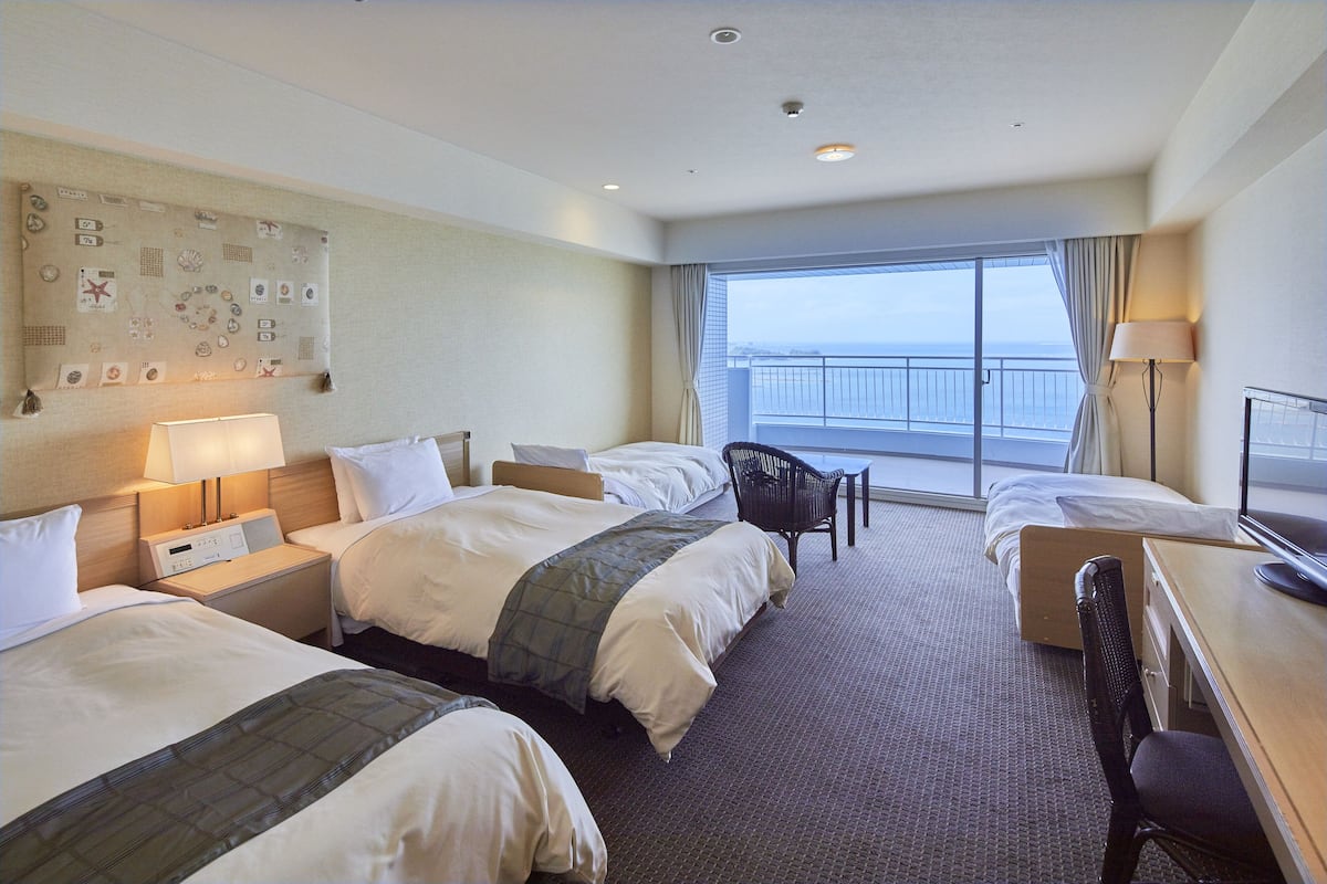 Habitación de hotel con dos camas, un escritorio, una silla, una lámpara y un ventanal con vistas al mar.