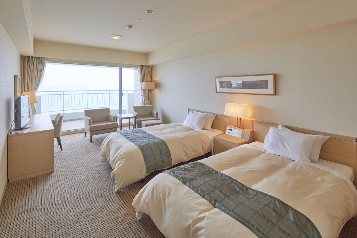 A hotel room with two beds, a desk, a chair, and a view of the sea.
