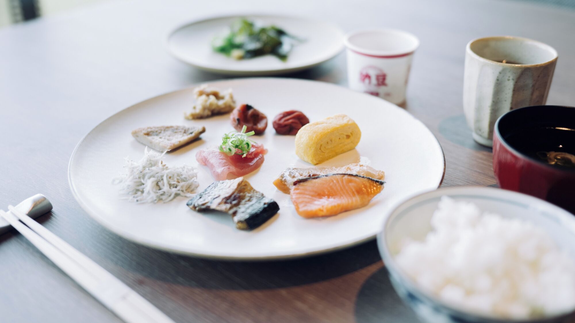 A plate with various food items, a bowl of rice, and a red cup on a table.