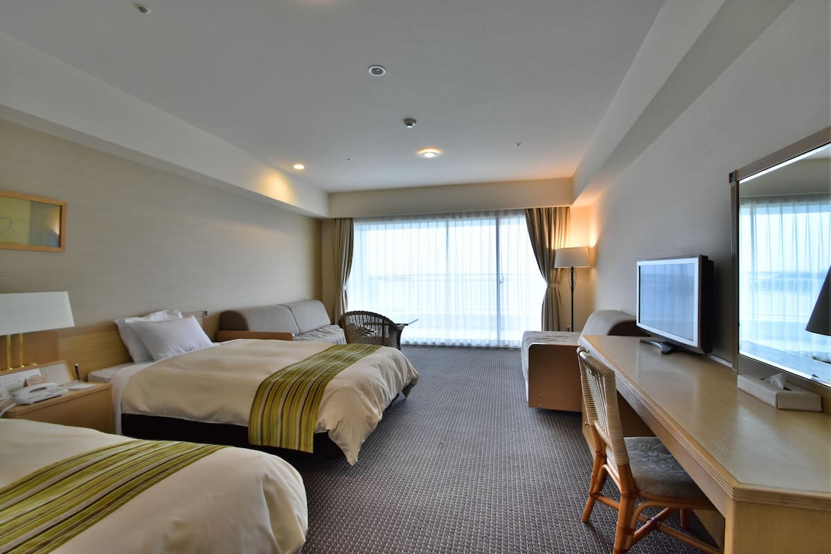 A hotel room with two beds, a desk with a television, a chair, and a large window with curtains.