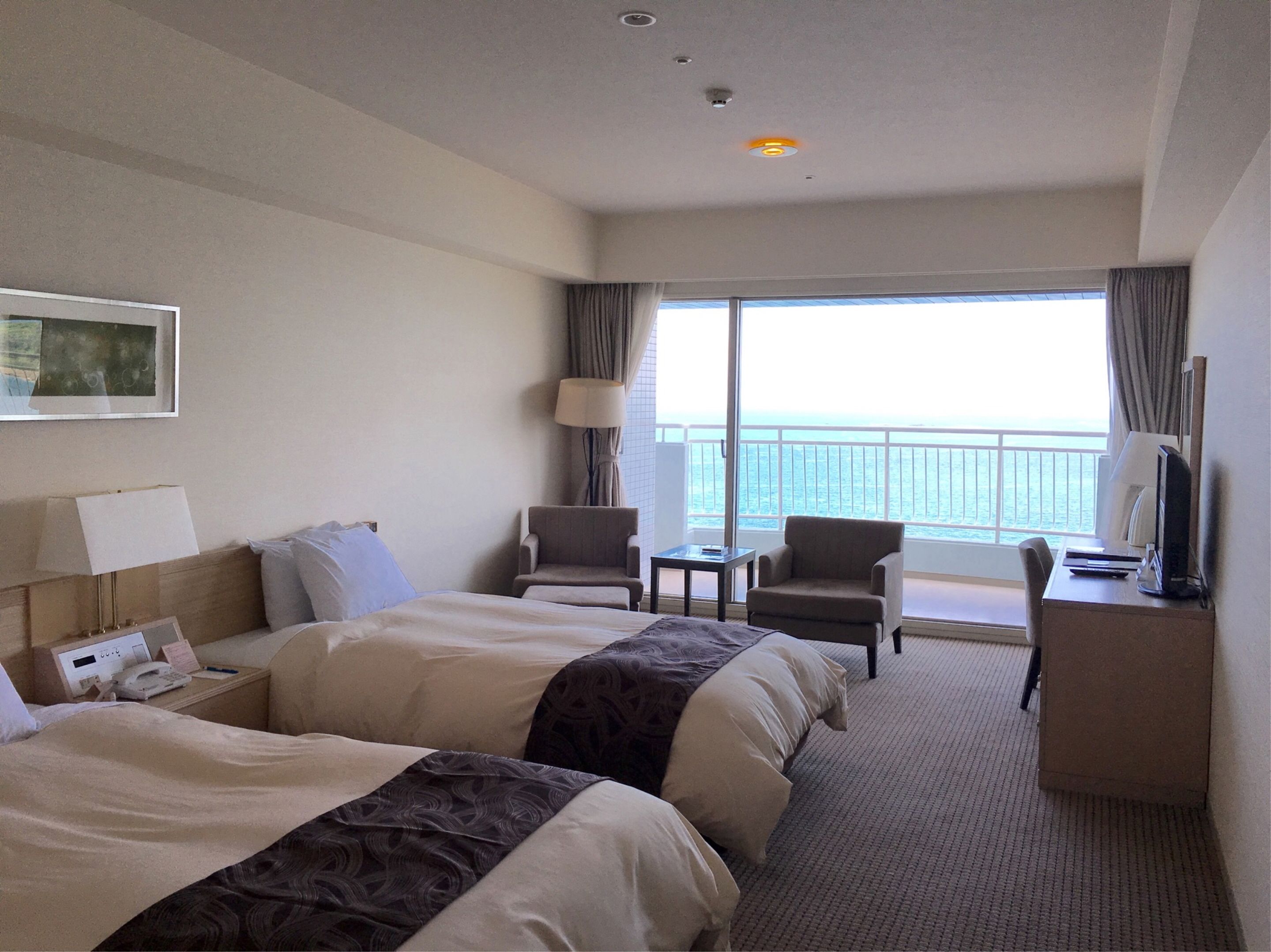 Habitación de hotel con dos camas, un escritorio, una silla, una lámpara y vista al mar.