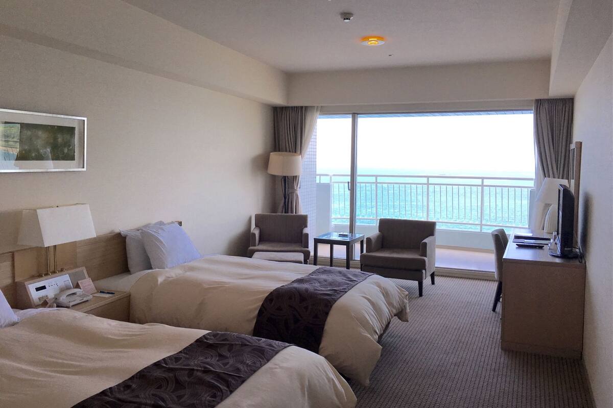 A hotel room with two beds, a desk, a chair, a lamp, and a view of the sea.