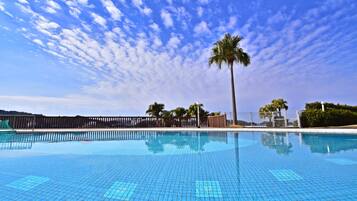 Piscina azul clara con borde de azulejos, rodeada por una valla y palmeras bajo un cielo parcialmente nublado.