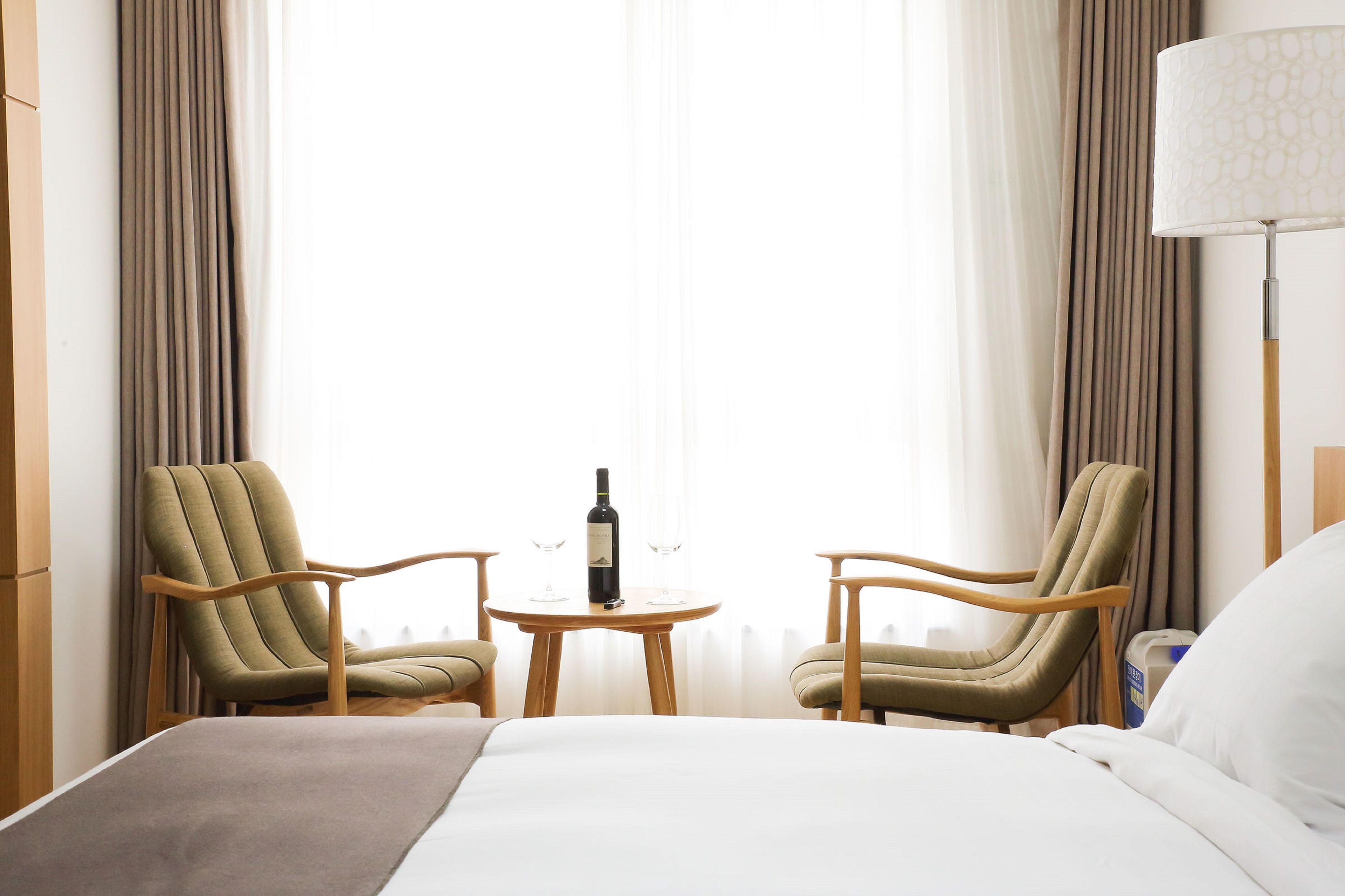 A hotel room with a bed, two armchairs, a small table with a bottle and glasses, and a lamp.
