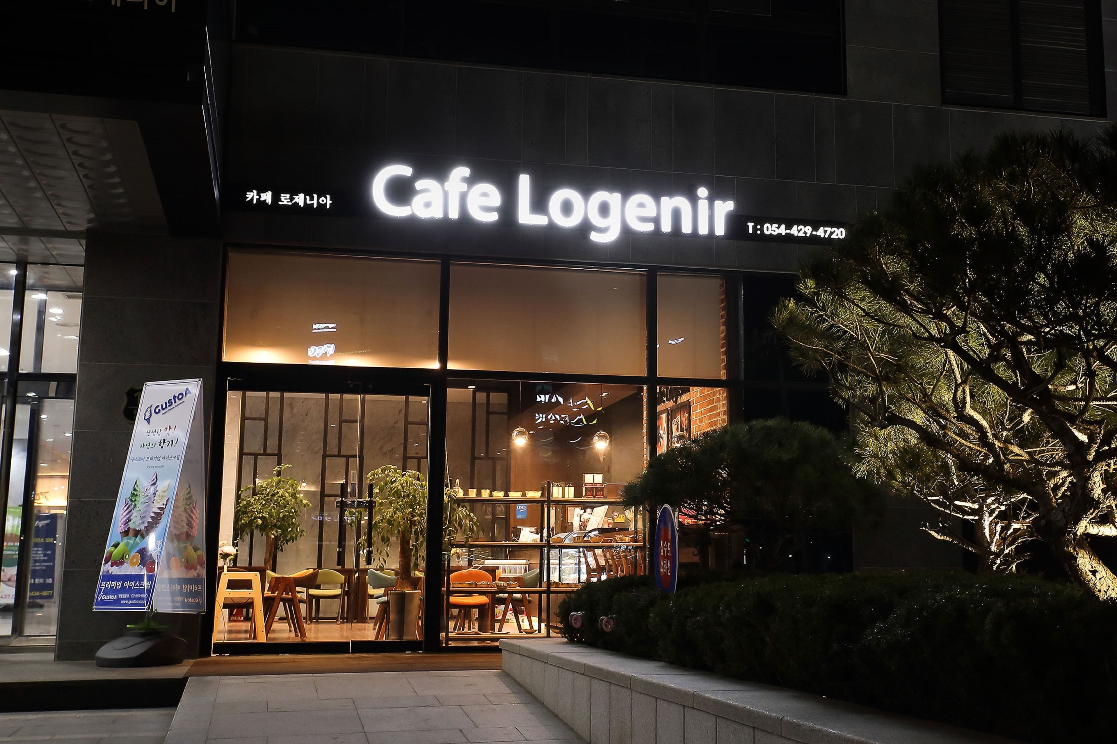 A Cafe Logenir storefront at night with illuminated signage, a glass entrance, and indoor seating visible through the windows.