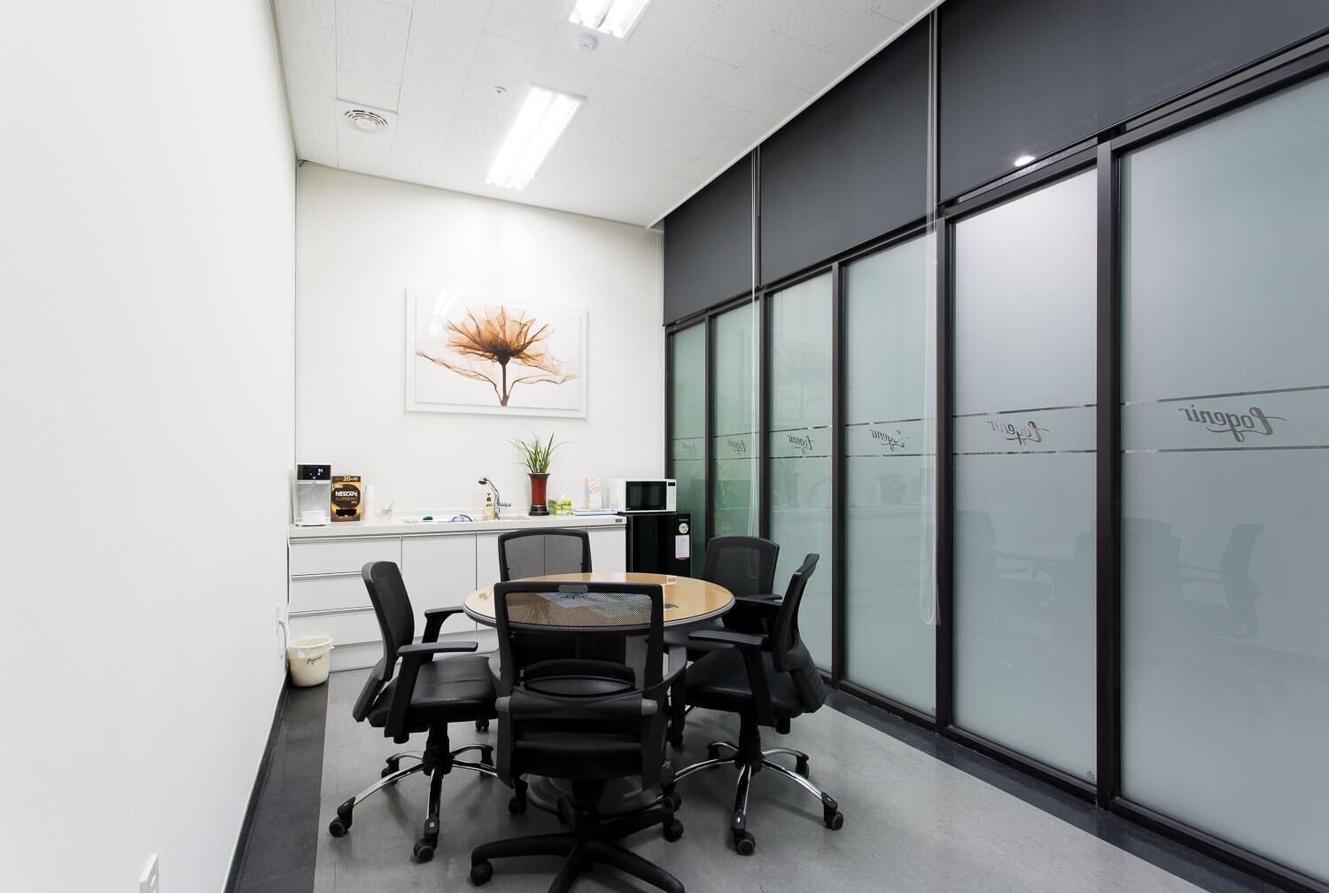 A modern office space with a round table, black office chairs, a whiteboard, and a large window with frosted glass.