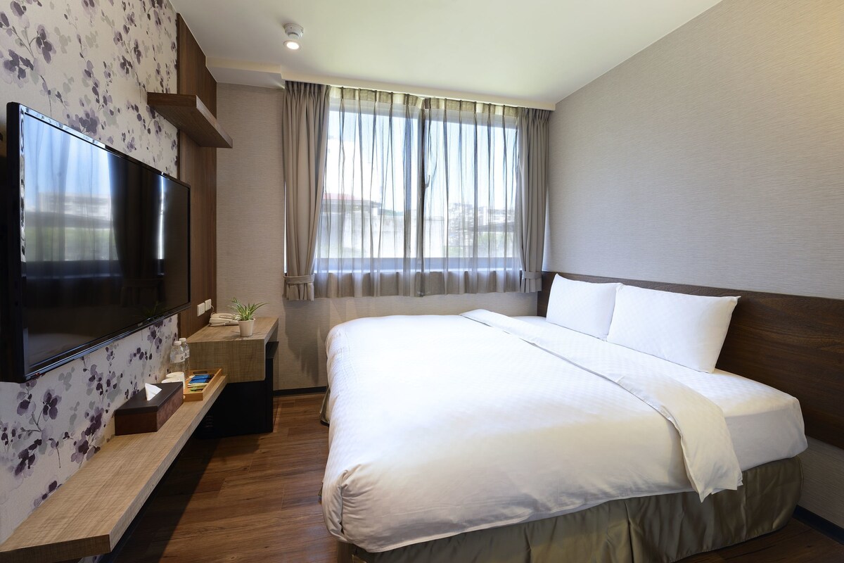 A hotel room with a large bed, a flat-screen TV, a wooden bedside table, a small desk with a chair, and a window with curtains.