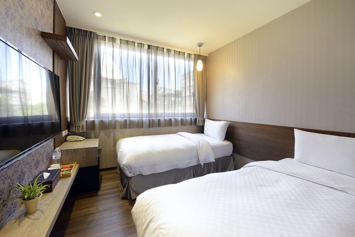A hotel room with two beds, a TV, a desk with a plant, and a window with curtains.