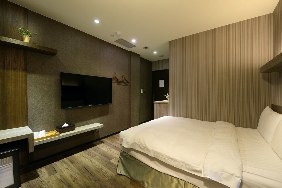 A modern hotel room with a large bed, a flat-screen TV, a wooden floor, and a small plant on a shelf.