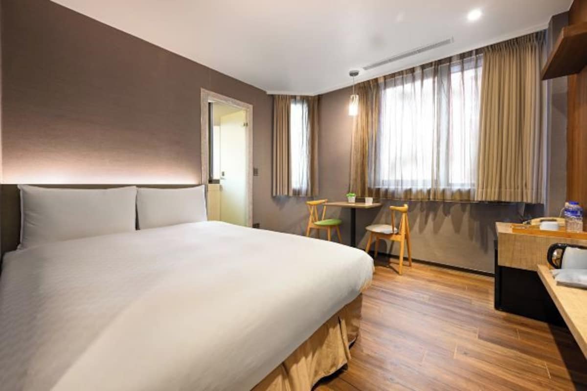 Ein Hotelzimmer mit einem großen Bett, einem Schreibtisch, zwei Stühlen und einem Fenster mit Vorhängen.