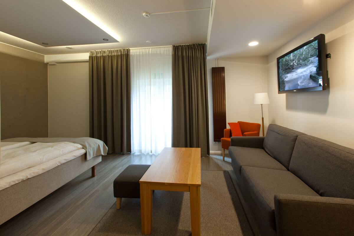 A modern hotel room with a bed, a sofa, a table, and a TV mounted on the wall.