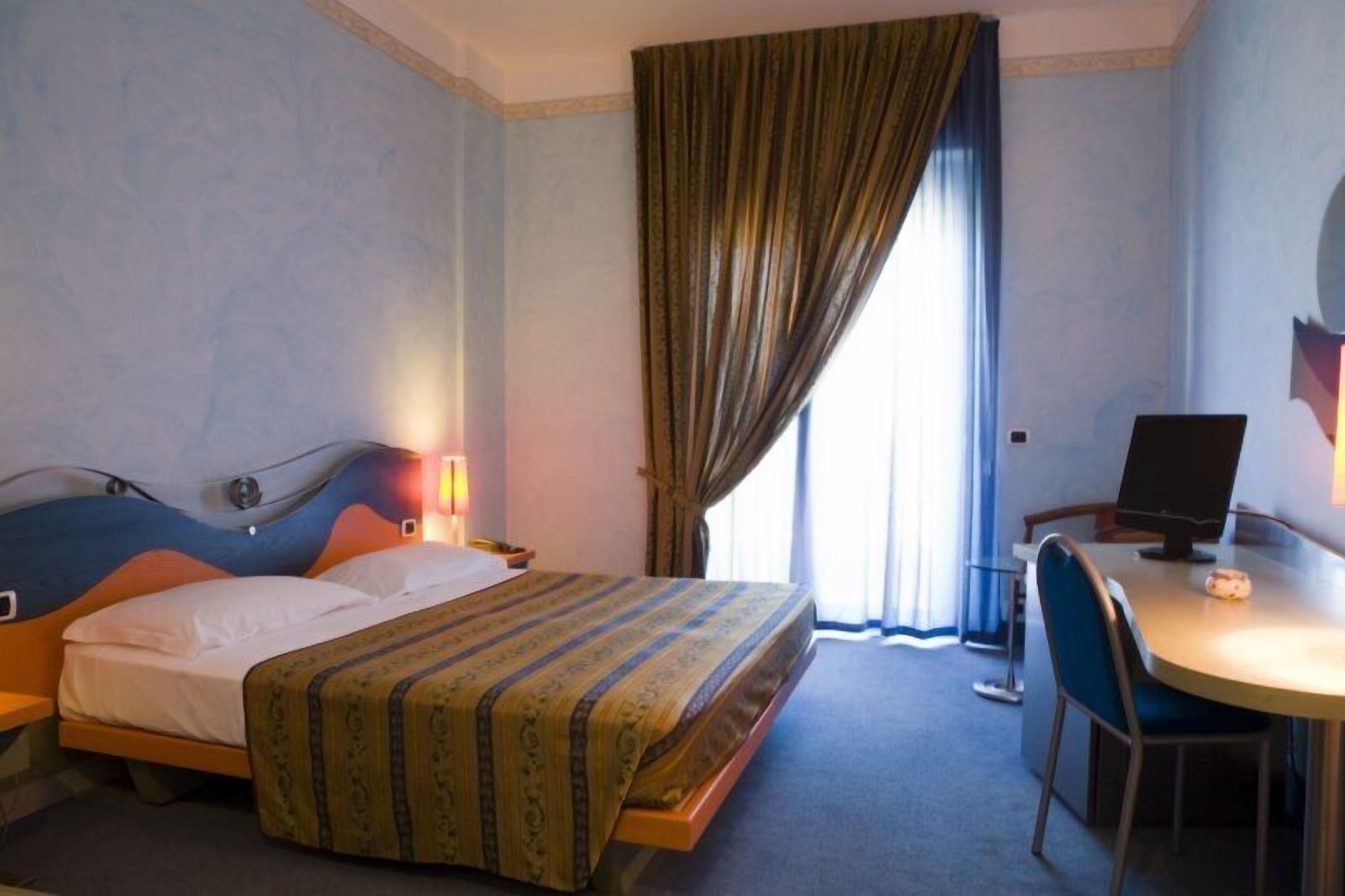 A hotel room with a bed, a desk, a chair, a lamp, and a window with curtains.