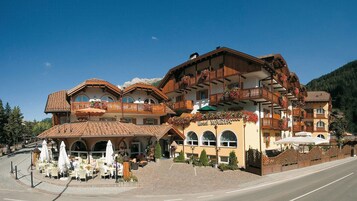 Un hotel de varios pisos con terraza al aire libre, balcones con jardineras y una vista despejada del paisaje montañoso circundante.