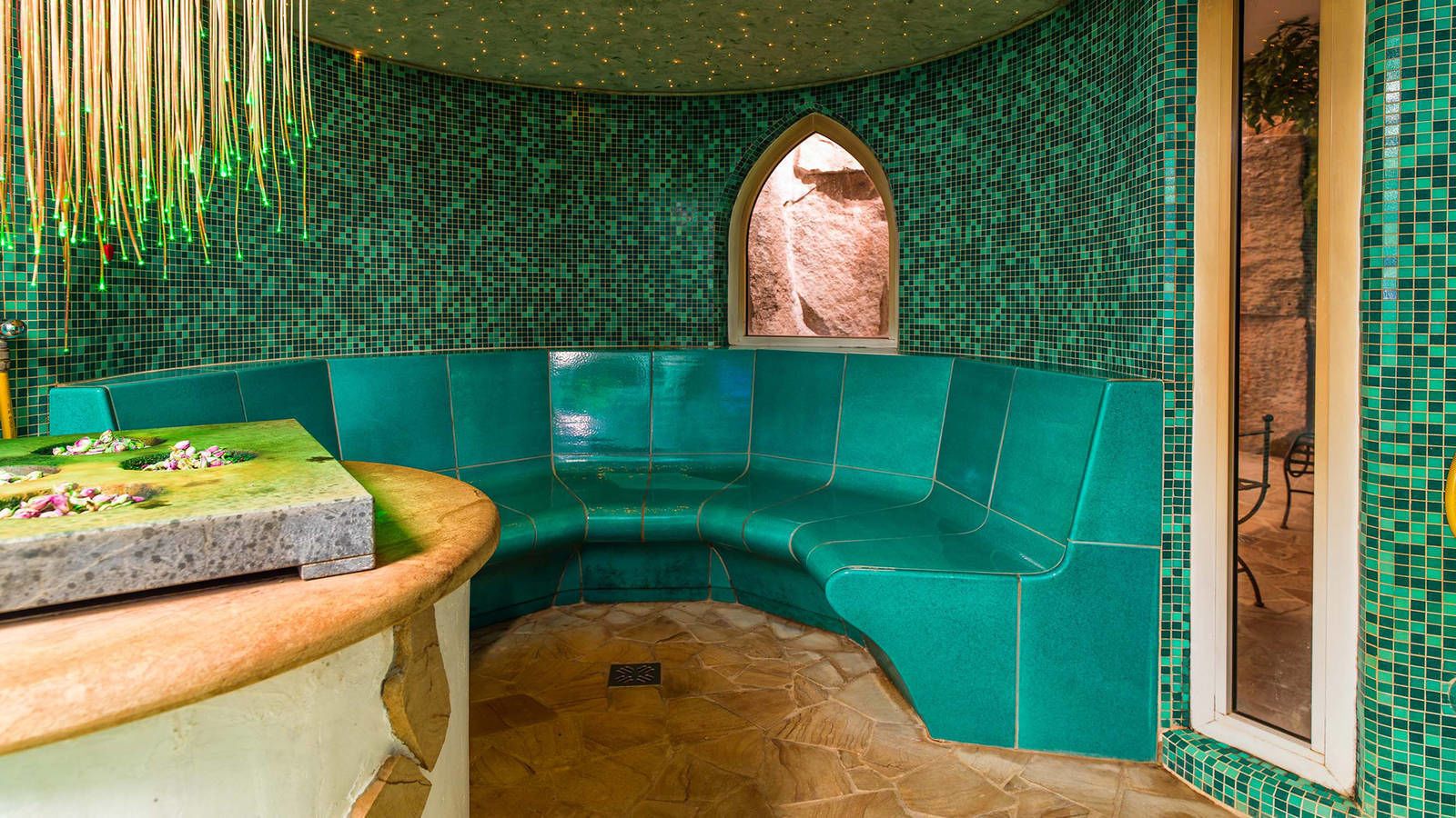 A corner room with teal mosaic walls, a stone floor, and a bench.