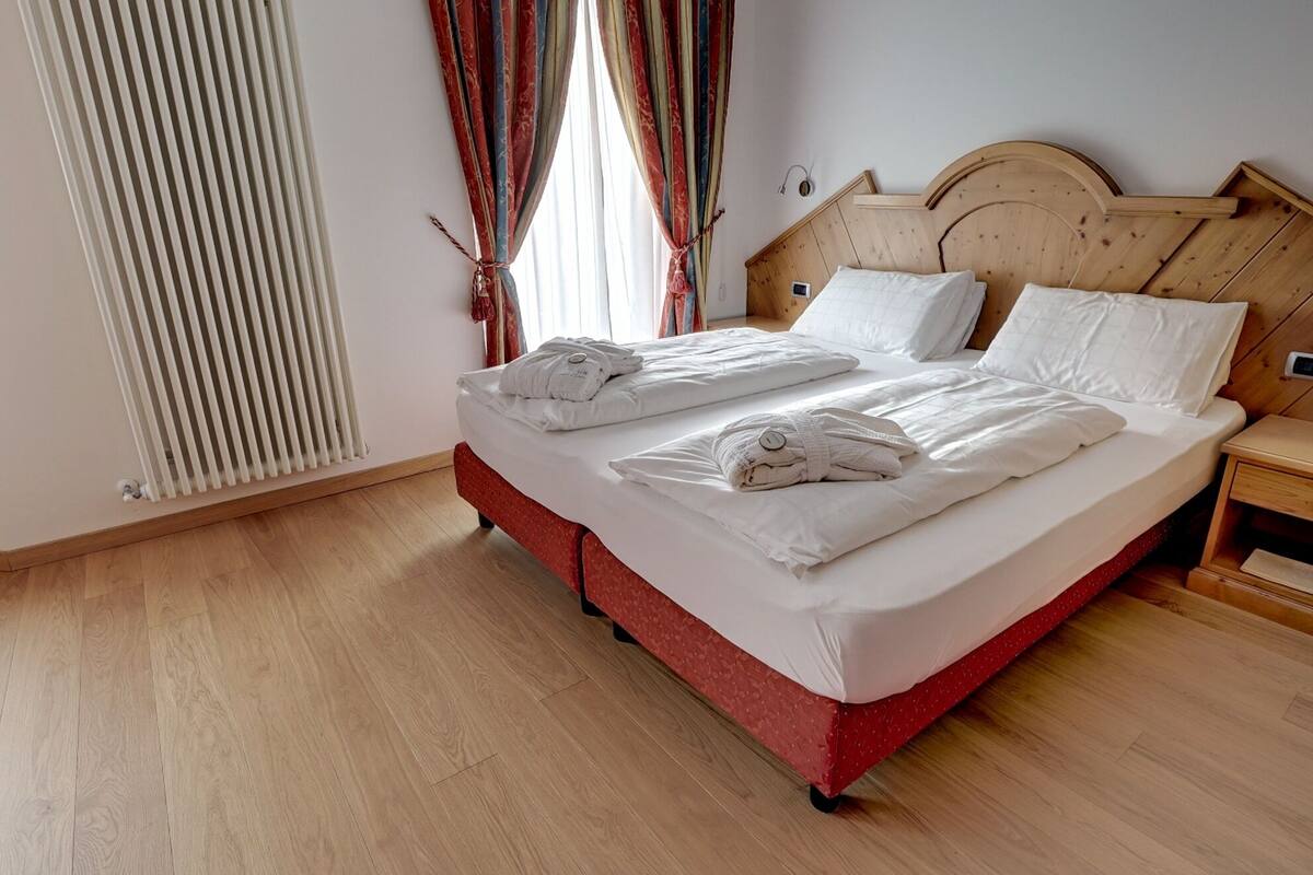 Cama doble con sábanas y toallas blancas, cabecera de madera y mesita de noche con lámpara.