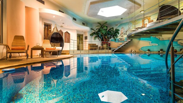 An indoor swimming pool with a tiled floor, a seating area with chairs, a staircase, and a large potted plant. - Leading Relax Hotel Maria (Moena)