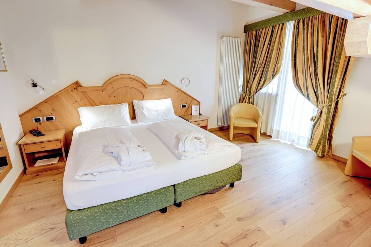 Una cama bien hecha con sábanas y toallas blancas, un cabecero de madera, una mesita de noche con lámpara, y una silla cerca de una ventana con cortinas.