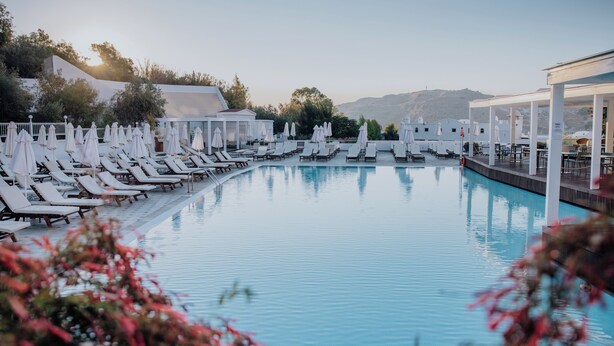 Un espace aménagé au bord de la piscine, avec des chaises longues et des parasols, entouré de bâtiments et de collines.