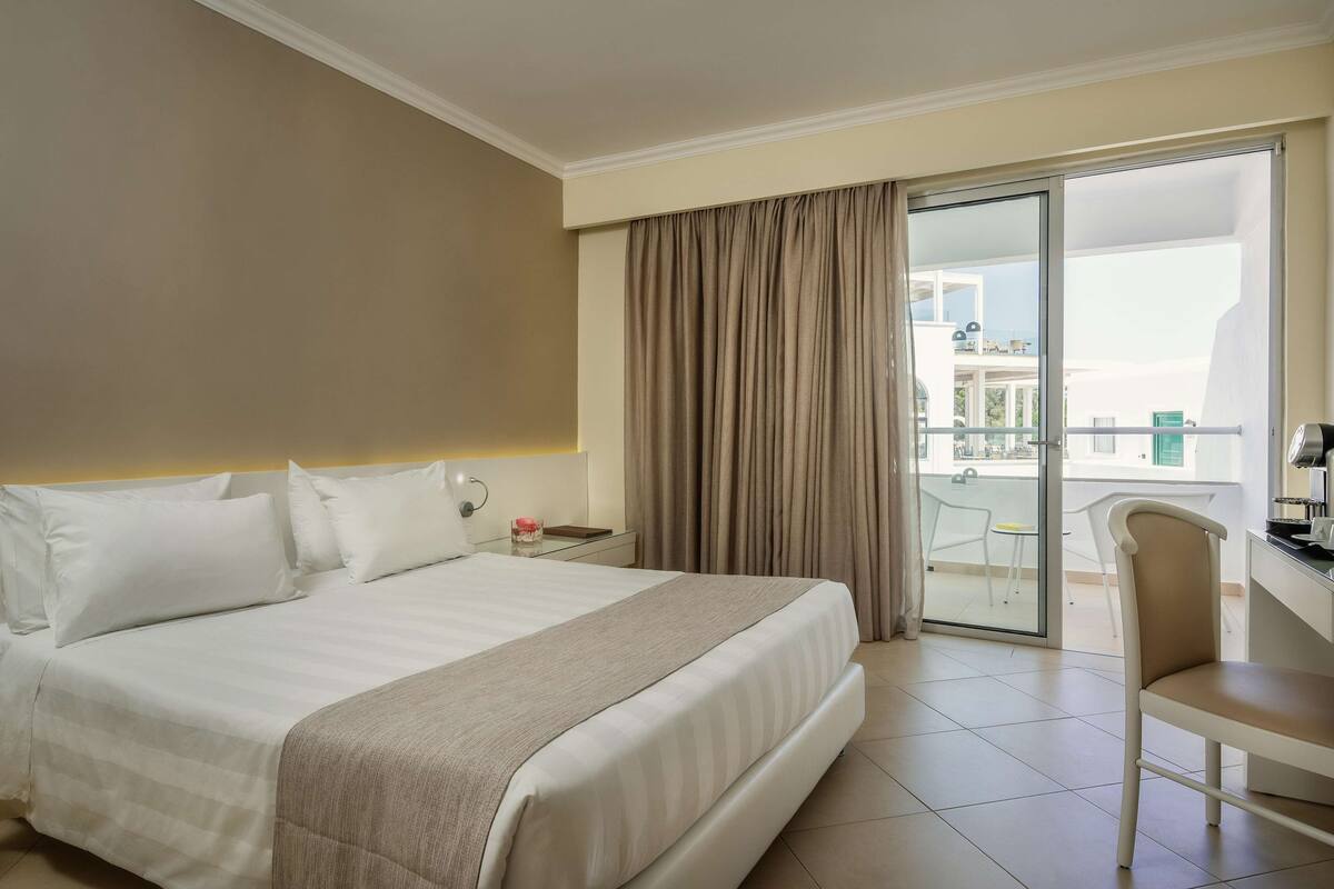 A hotel room with a bed, a desk, a chair, a nightstand, and a view of the outside balcony.