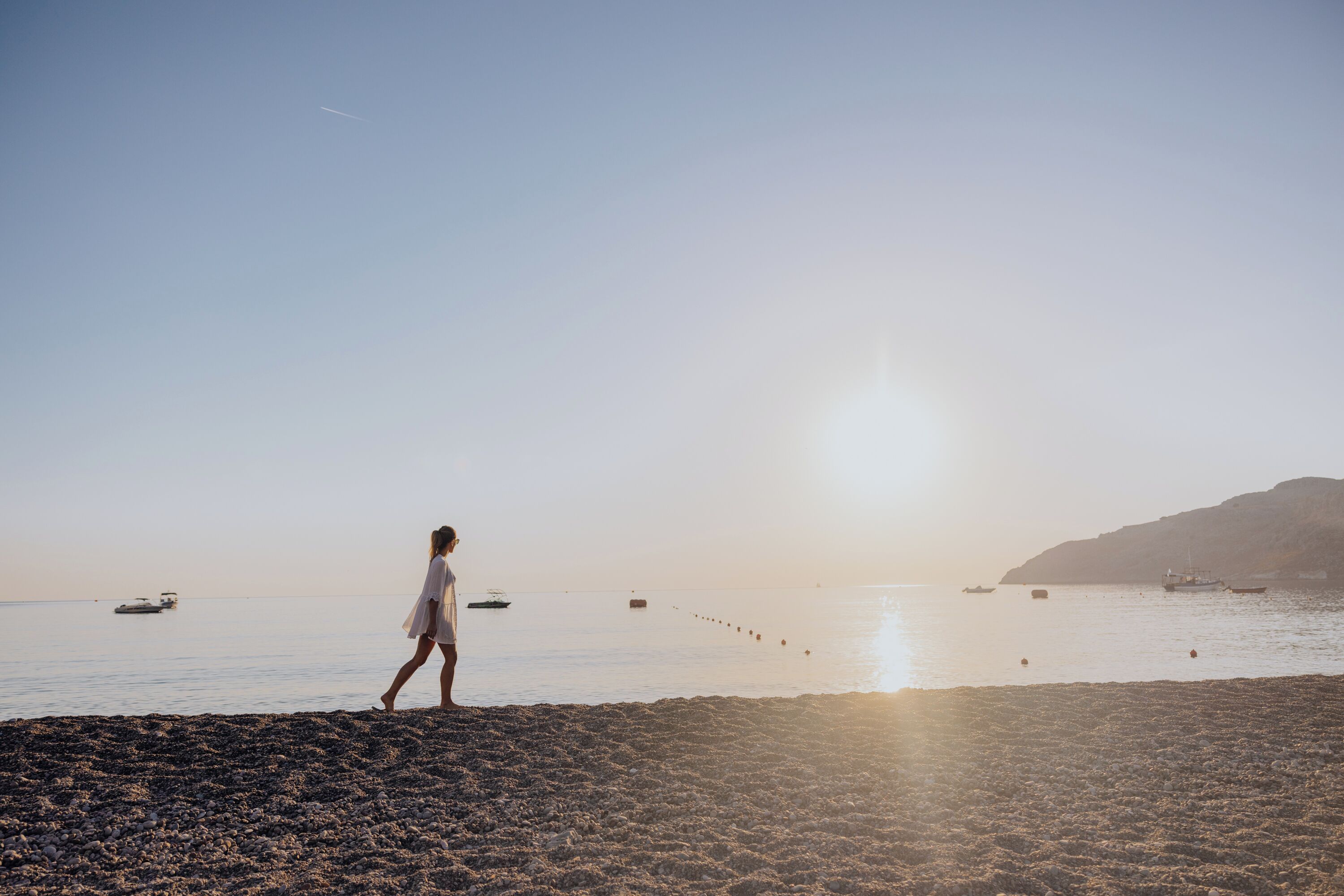 A beach scene with a person walking along the shore, boats in the water, and a clear sky.