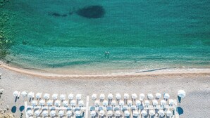 Une vue aérienne d’une plage aux eaux turquoise et limpides, bordée d’un rivage sablonneux et ponctuée de rangées de parasols blancs.