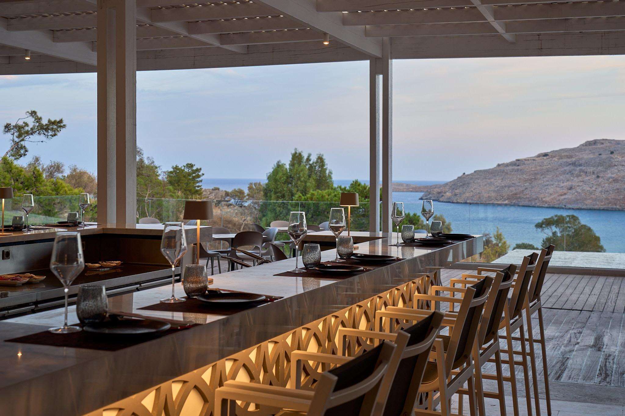 A modern outdoor dining area with a long table, chairs, and a view of the sea and hills.