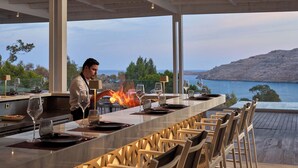 A bar with a long counter, outdoor seating, and a view of the sea. - Lindos Village Resort & Spa - Adults Only (Rhodes)