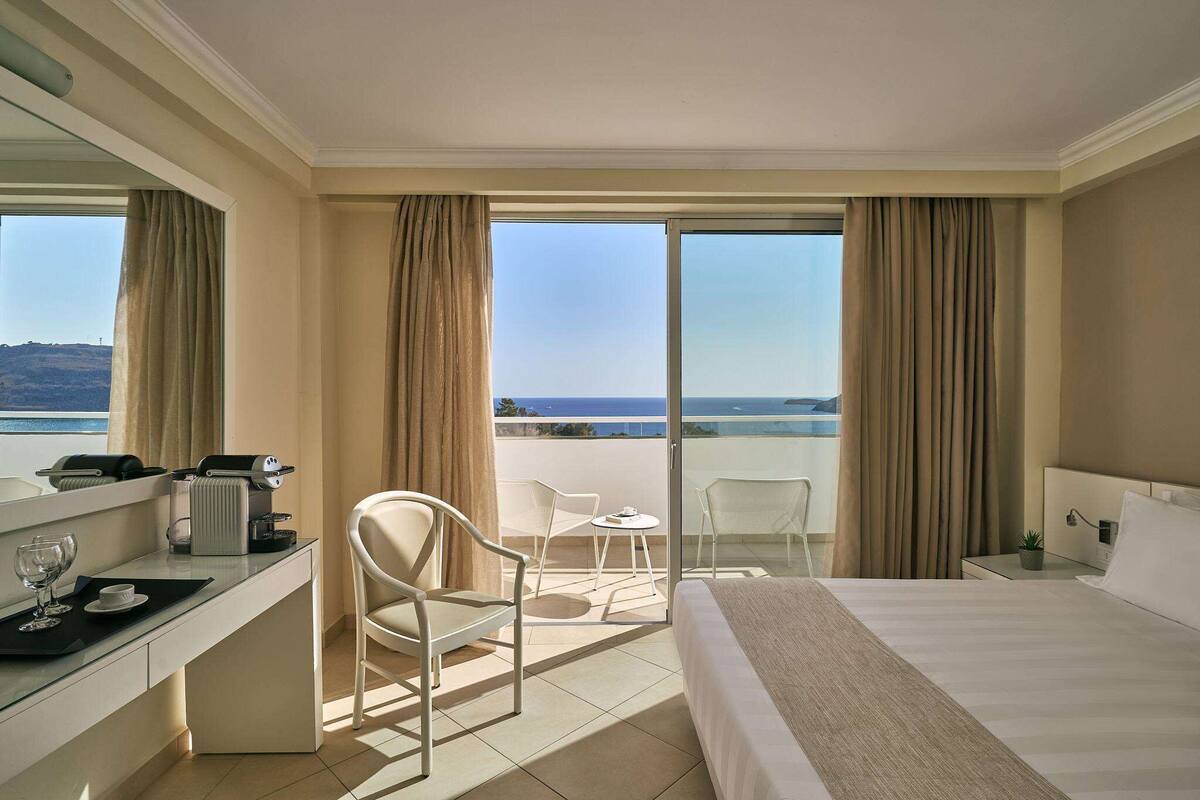A hotel room with a bed, a desk with a coffee machine, a chair, a small table with a cup, and a view of the sea through large windows.